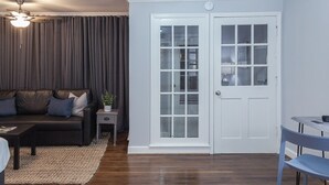 Un salon avec un canapé en cuir foncé, une petite table d’appoint, une table basse et un ventilateur de plafond. On y trouve une porte blanche avec des panneaux de verre et une chaise bleue.