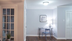 Une pièce avec une porte, une fenêtre avec des stores, une table avec une chaise et une lampe fixée au mur.