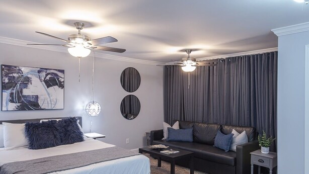 Une chambre moderne avec un lit, deux ventilateurs de plafond, un canapé de couleur sombre, une petite table et un grand tableau abstrait accroché au mur.