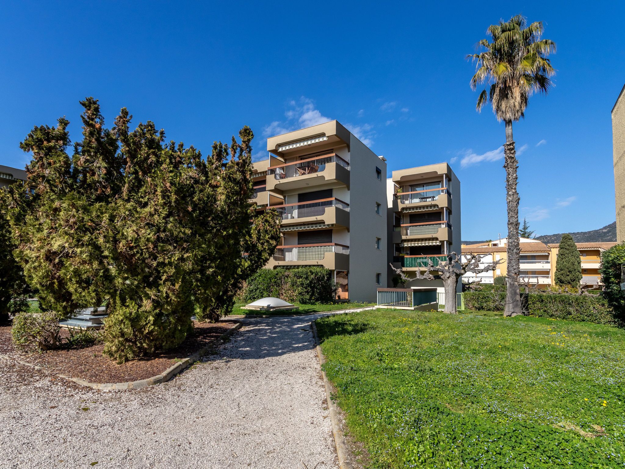 A multi-story residential building with balconies, surrounded by greenery and a clear sky.