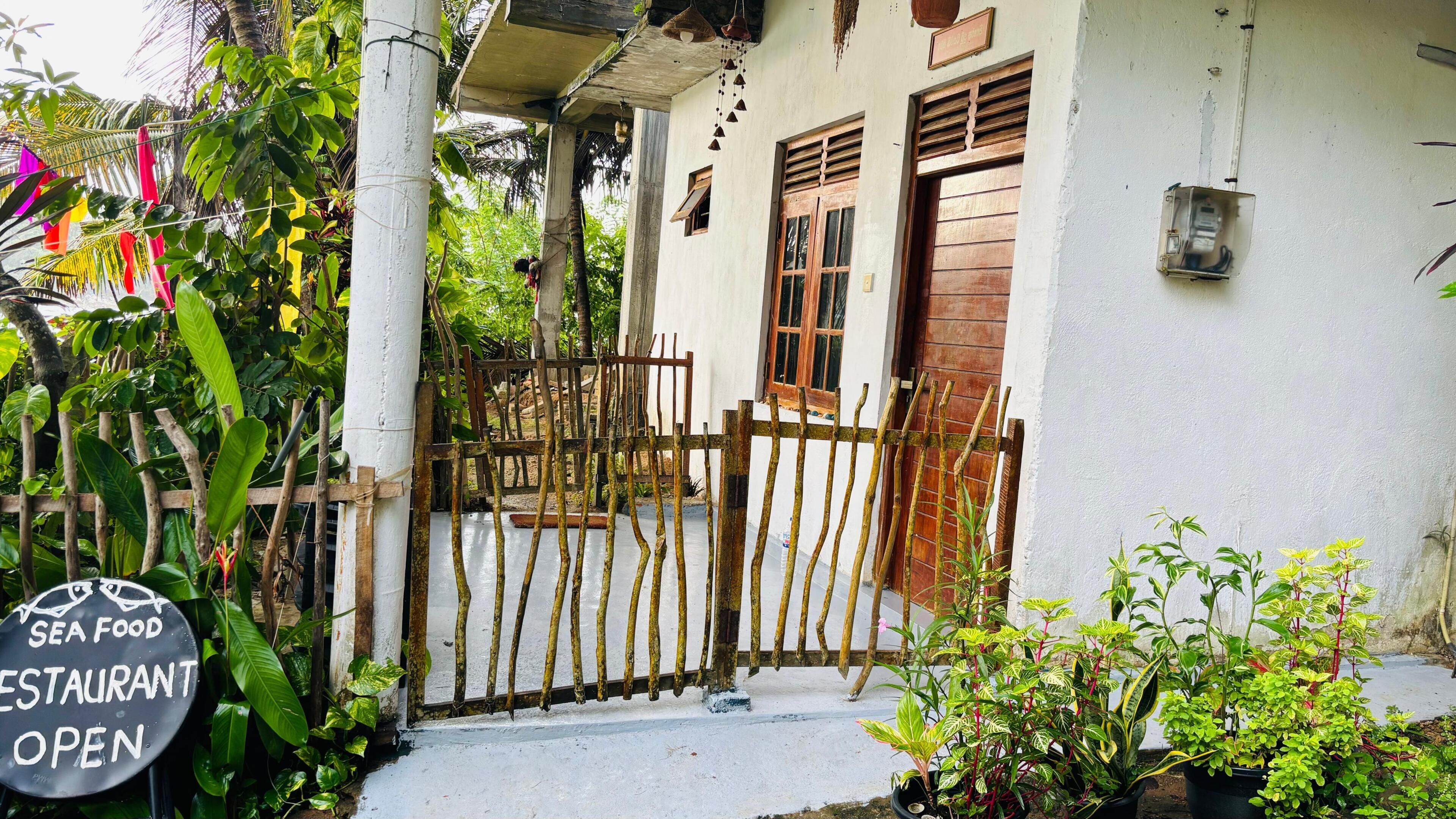Un bâtiment blanc avec une porte en bois, une enseigne indiquant qu’un restaurant est ouvert, et une végétation luxuriante tout autour.