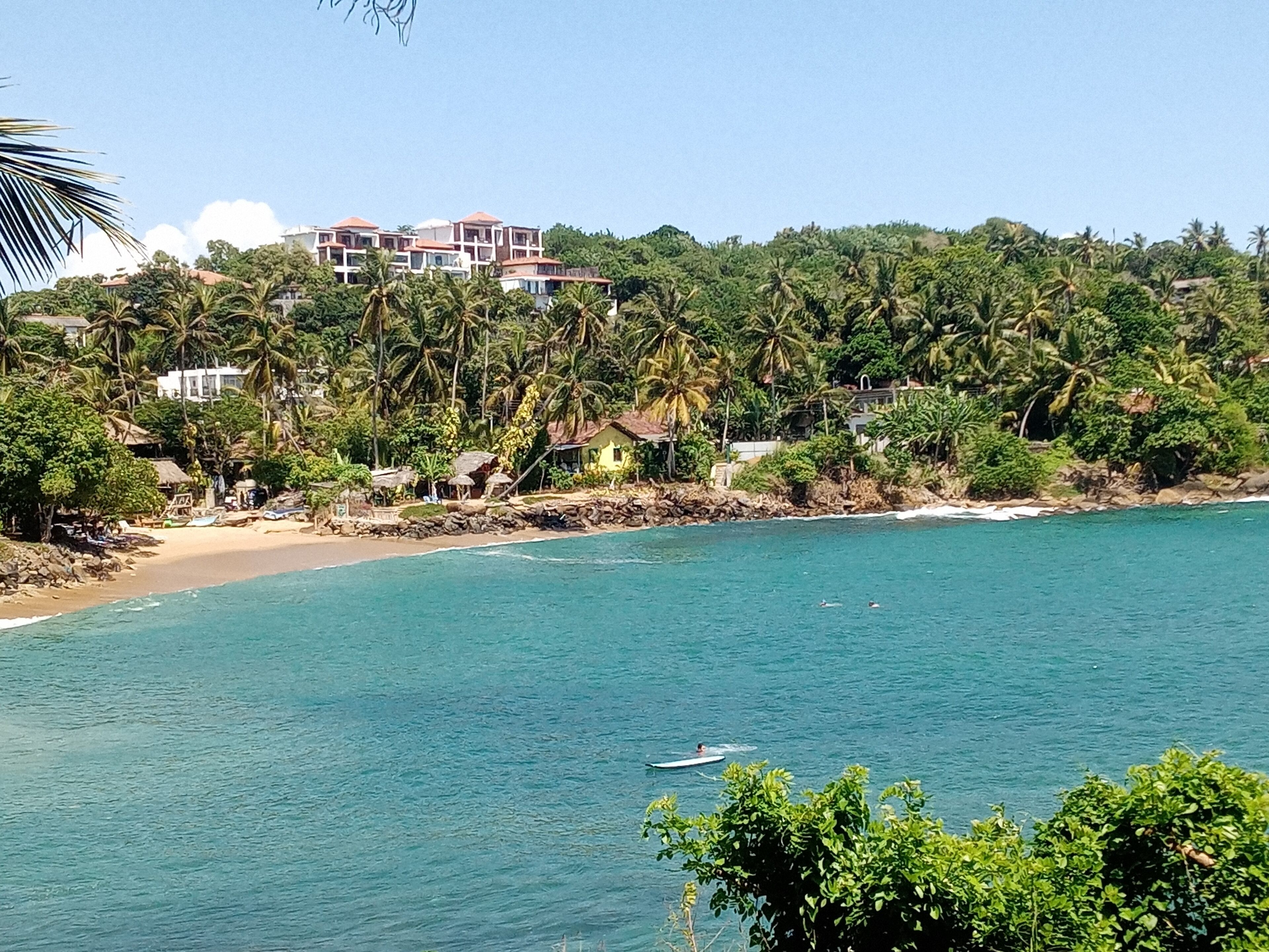 A coastal area with a beach, a building with multiple balconies overlooking the sea, and dense tropical vegetation.