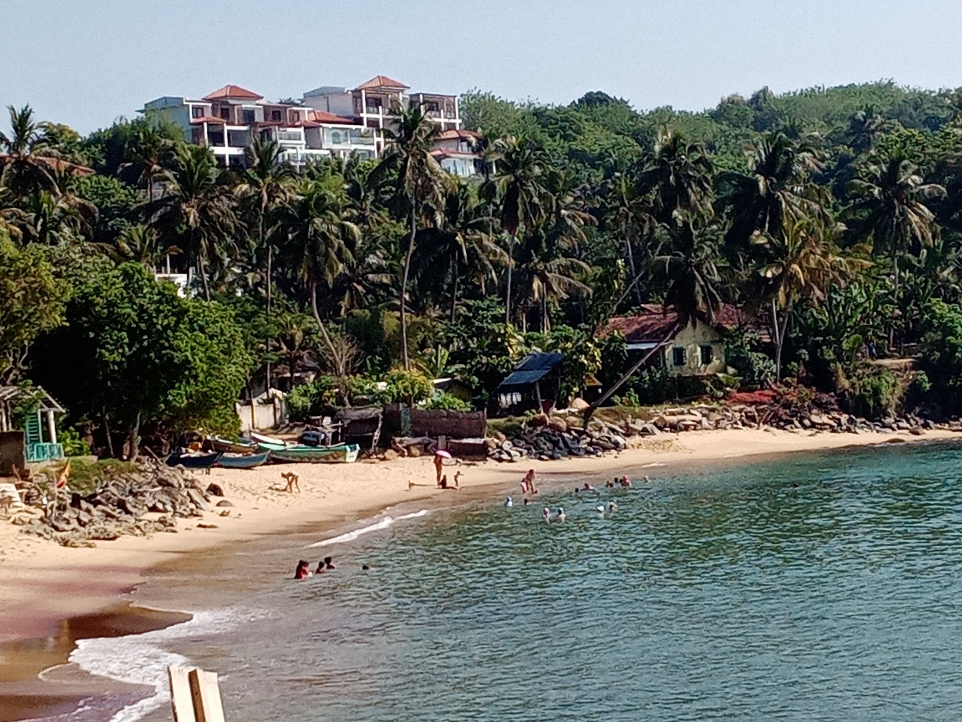 Ein Strand mit badenden Personen und einigen Booten, umgeben von Palmen und Gebäuden im Hintergrund.