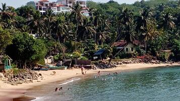 Ein Strand mit badenden Personen und einigen Booten, umgeben von Palmen und Gebäuden im Hintergrund.