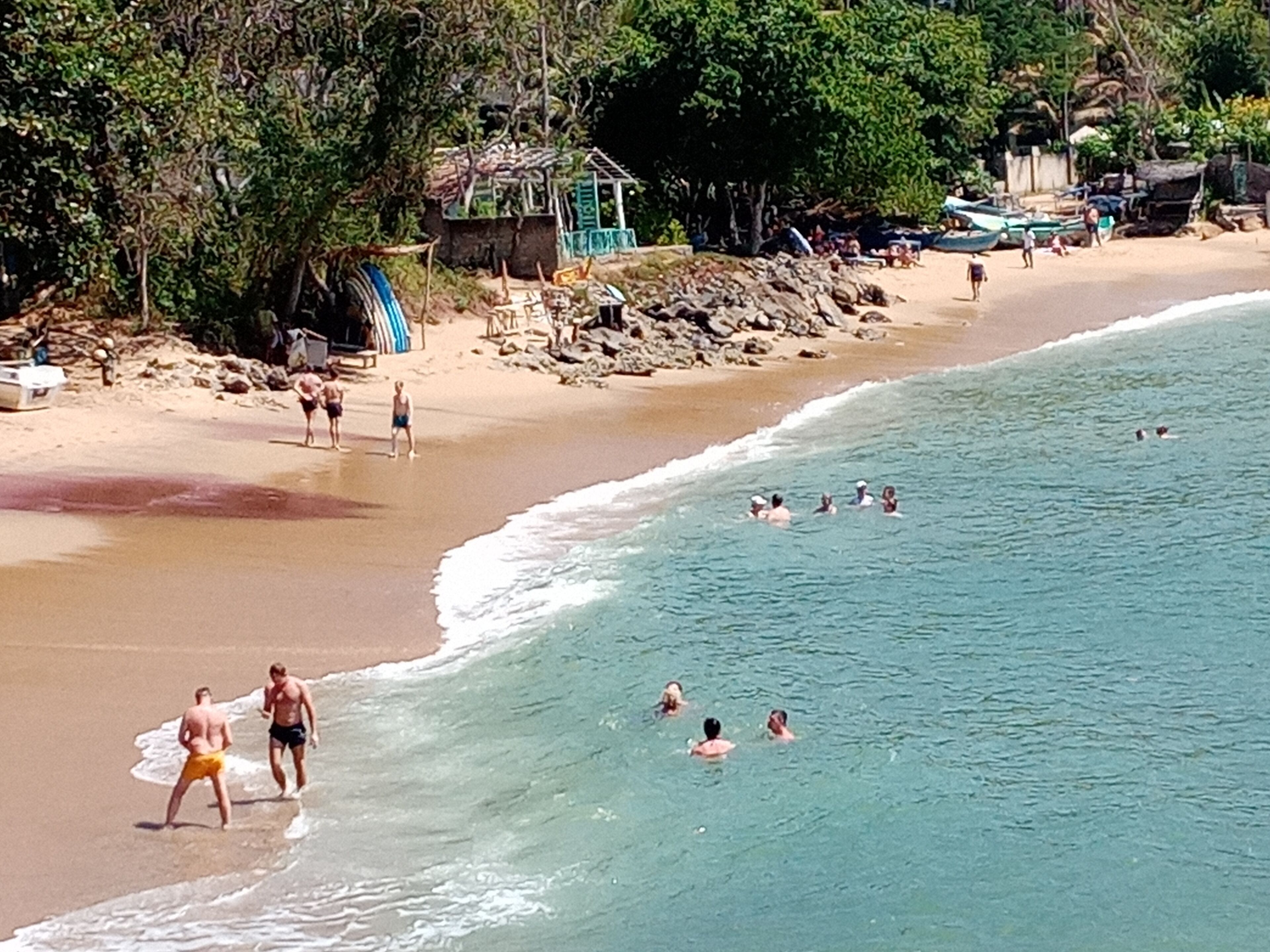 Uma praia com pessoas nadando e caminhando pela orla. Há barcos e uma pequena construção na praia.