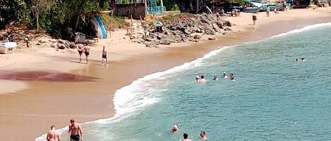 Uma praia com pessoas nadando e caminhando pela orla. Há barcos e uma pequena construção na praia.