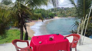 Una mesa con mantel rojo, dos sillas y un centro de mesa decorativo en un balcón con vista a una playa con palmeras y un cielo despejado.