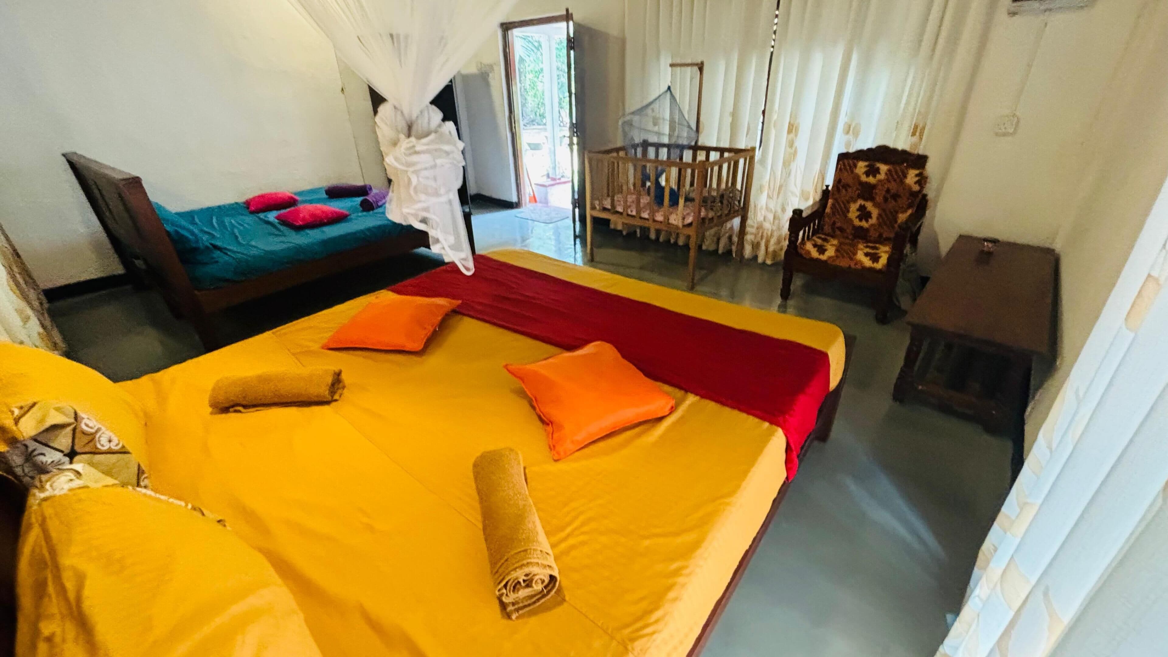 Une chambre avec deux lits, l’un recouvert d’une couvre-lit jaune et l’autre d’une couvre-lit verte. On y trouve une chaise avec un coussin à motifs et une petite table.