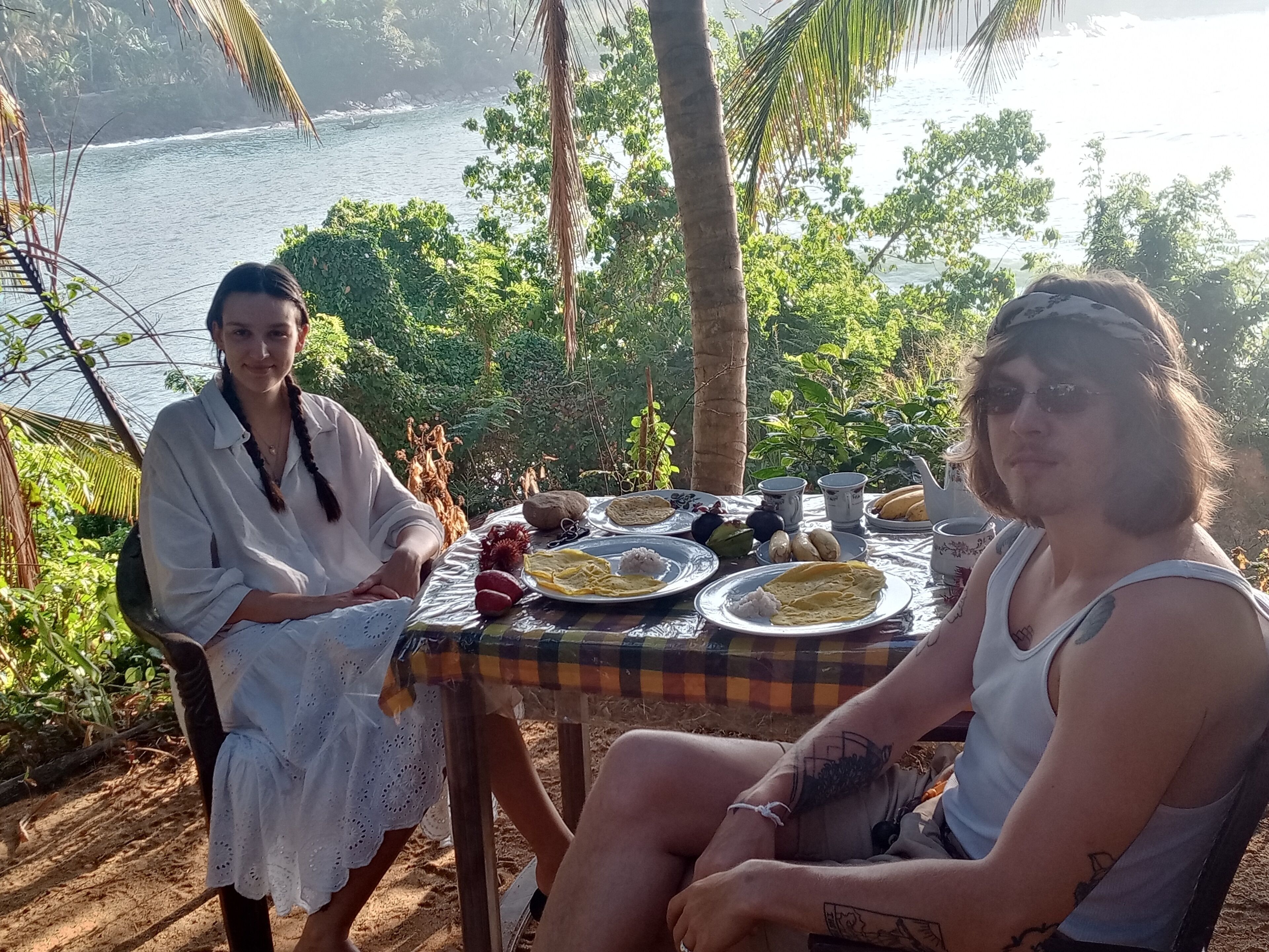 Two people seated at an outdoor table with a view of the sea, surrounded by greenery.