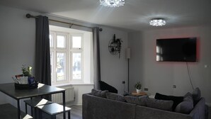 A modern living room with a grey sofa, a dining table with chairs, a flat-screen TV mounted on the wall, and a large window with curtains. - Lovely 3-Bed House in Solihull (Solihull)