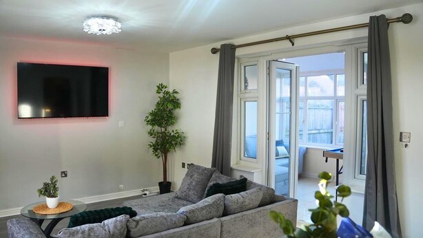 A modern living room with a grey sofa, a flat-screen TV mounted on the wall, a potted plant, and a view of the outside balcony. - Lovely 3-Bed House in Solihull (Solihull)
