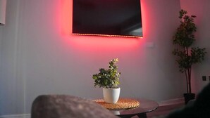 A modern living room with a flat-screen TV mounted on the wall, surrounded by a pink ambient light. There is a potted plant on a woven table in the foreground. - Lovely 3-Bed House in Solihull (Solihull)