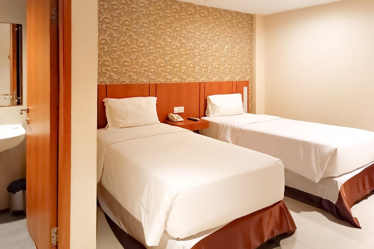 A hotel room with two beds, a wooden headboard, a wall with a floral pattern, a bedside table with a phone, and a bathroom visible through an open door.