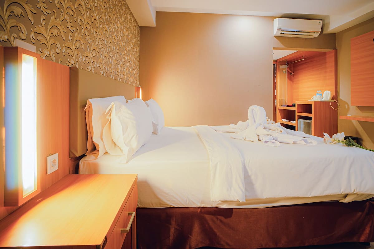 A hotel room with a neatly made bed, a wooden nightstand, a built-in wardrobe, and an air conditioning unit on the wall.