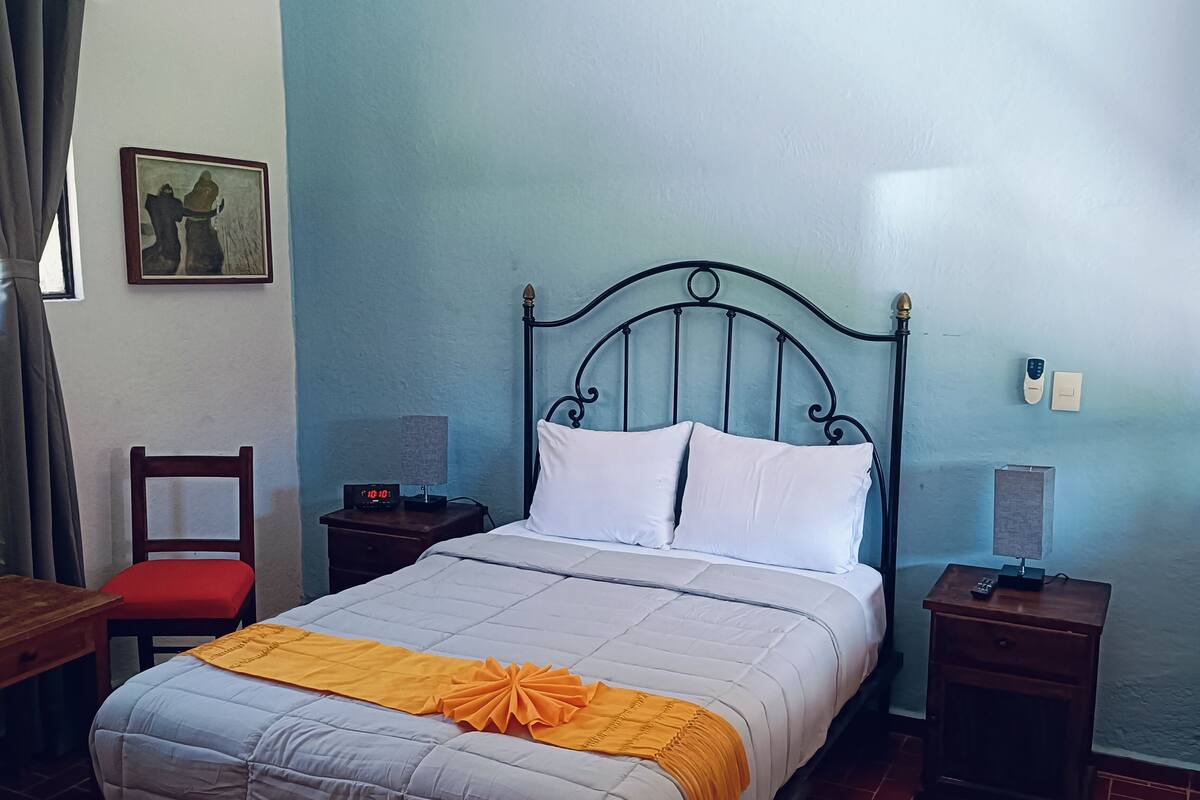 A neatly made bed with white and gray bedding, a wooden nightstand with a lamp, a chair, and a picture on the wall.