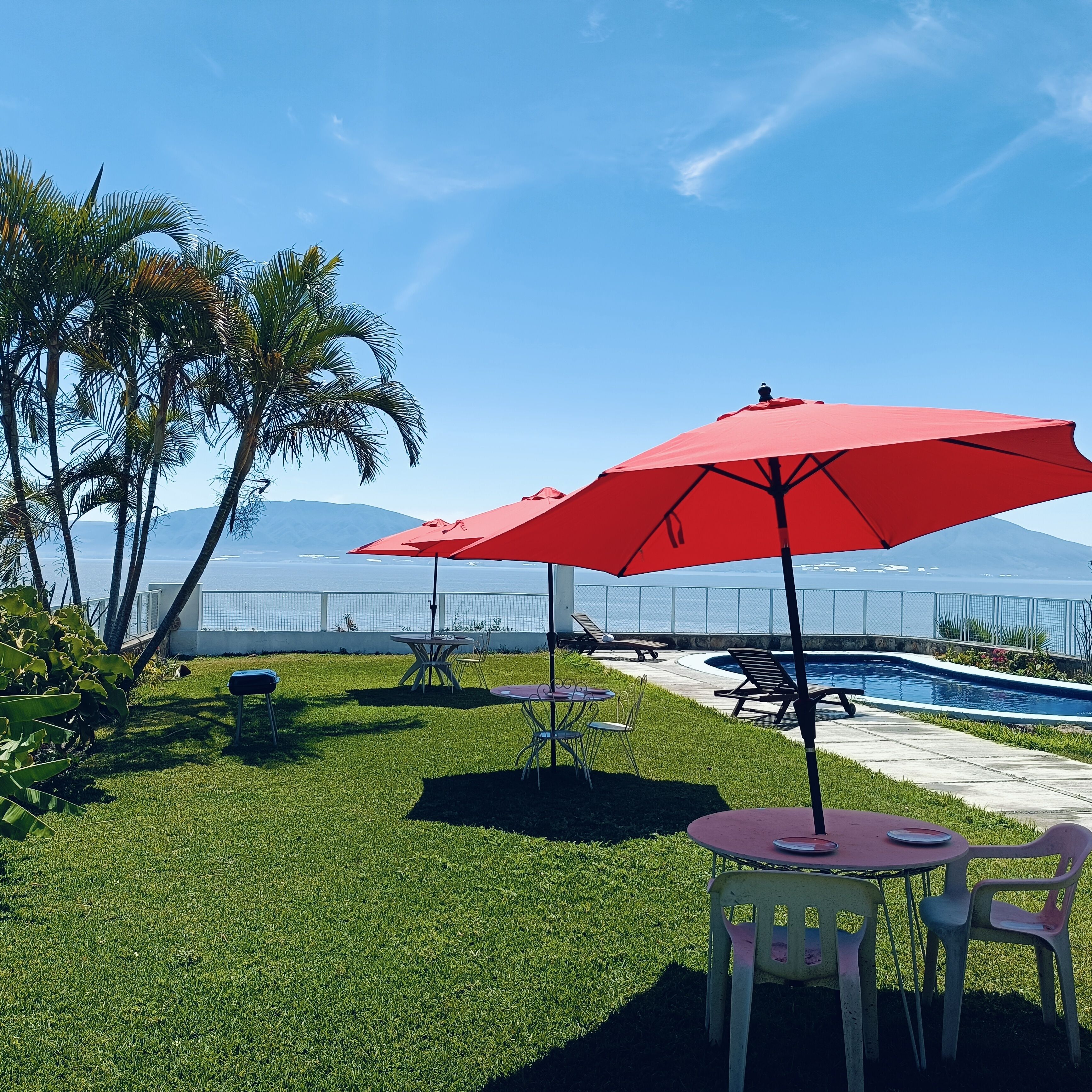 Zona de piscina con sombrillas rojas, una piscina y vista al océano.