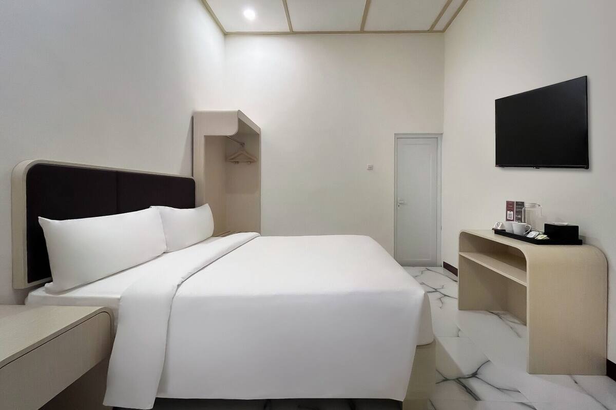 A modern hotel room with a neatly made bed, a bedside table, a built-in desk with a chair, a flat-screen TV mounted on the wall, and a door leading to another room.