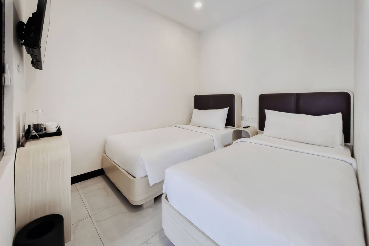 A hotel room with two beds, a bedside table, a small table with a cup and a book, and a wall-mounted TV.