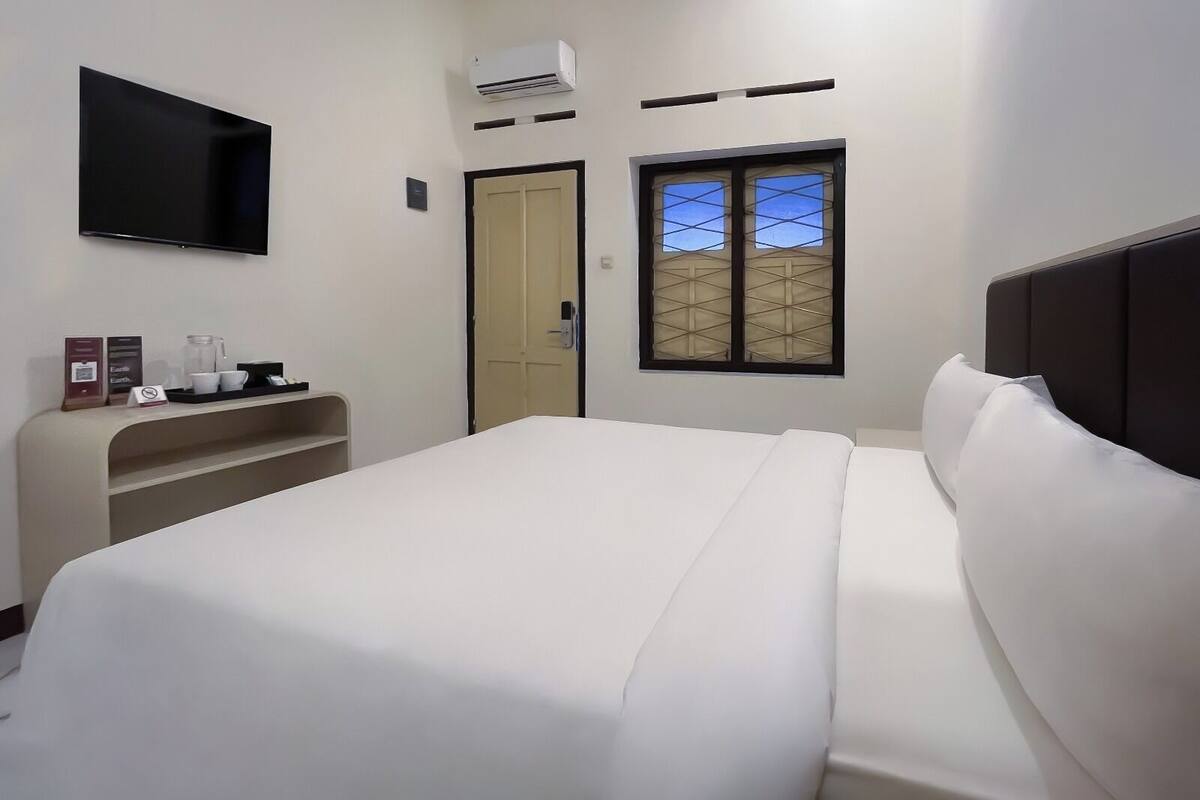 A hotel room with a bed, a TV mounted on the wall, an air conditioning unit, a window with a view, and a small shelf with a coffee maker and cups.