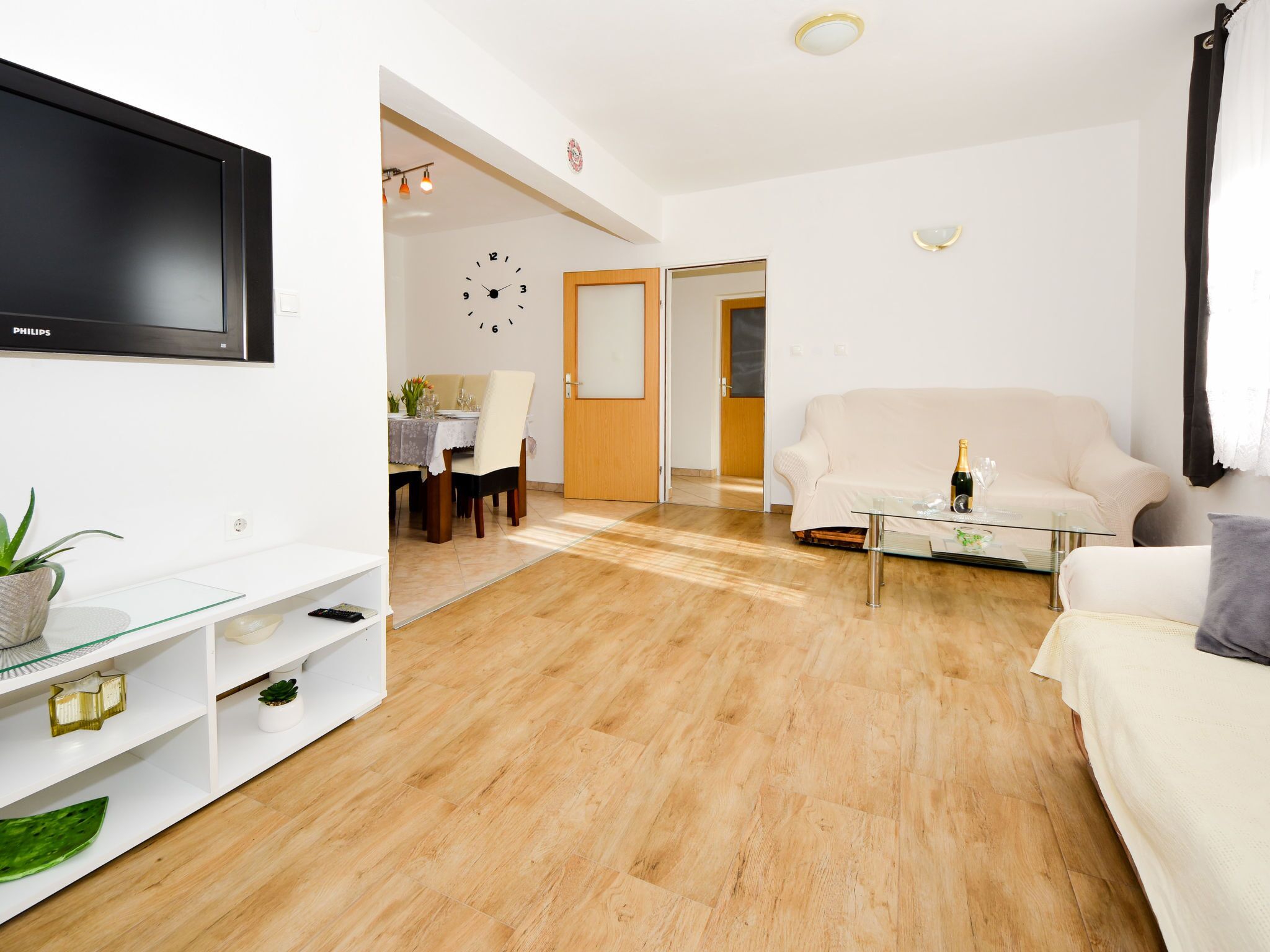 A modern living room with a flat-screen TV, a glass coffee table, a beige sofa, and a dining area with a table and chairs.