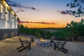 Un foyer en pierre, entouré de gravier, avec quatre chaises Adirondack, offrant une vue sur un paysage boisé et un bâtiment en arrière-plan.