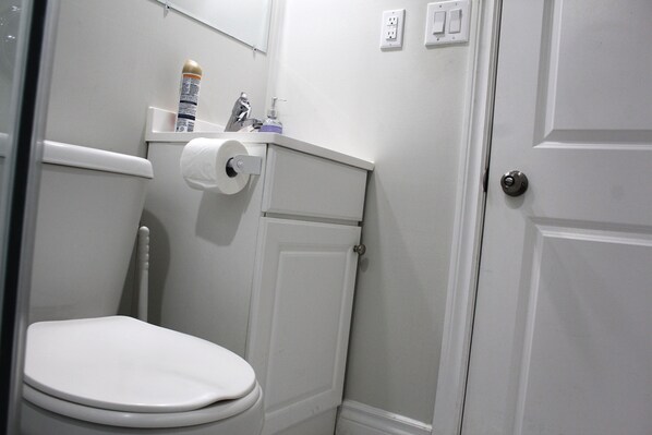 Une petite salle de bain blanche, avec des toilettes, un rouleau de papier toilette et un flacon de savon pour les mains posé sur le lavabo.