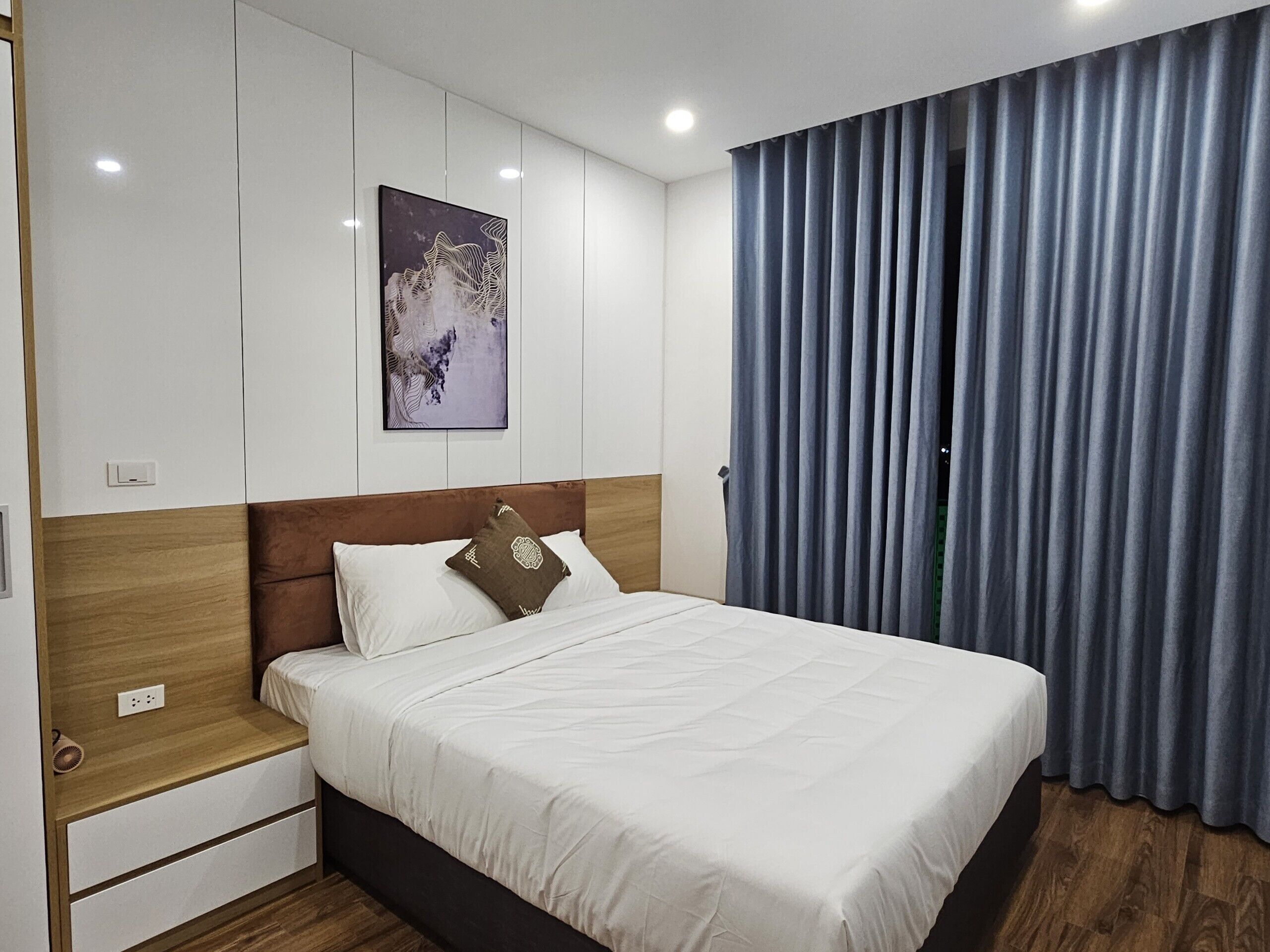 Ein ordentlich eingerichtetes Schlafzimmer mit Bett, Nachttisch, Wandbild und einem großen Fenster mit Vorhängen.