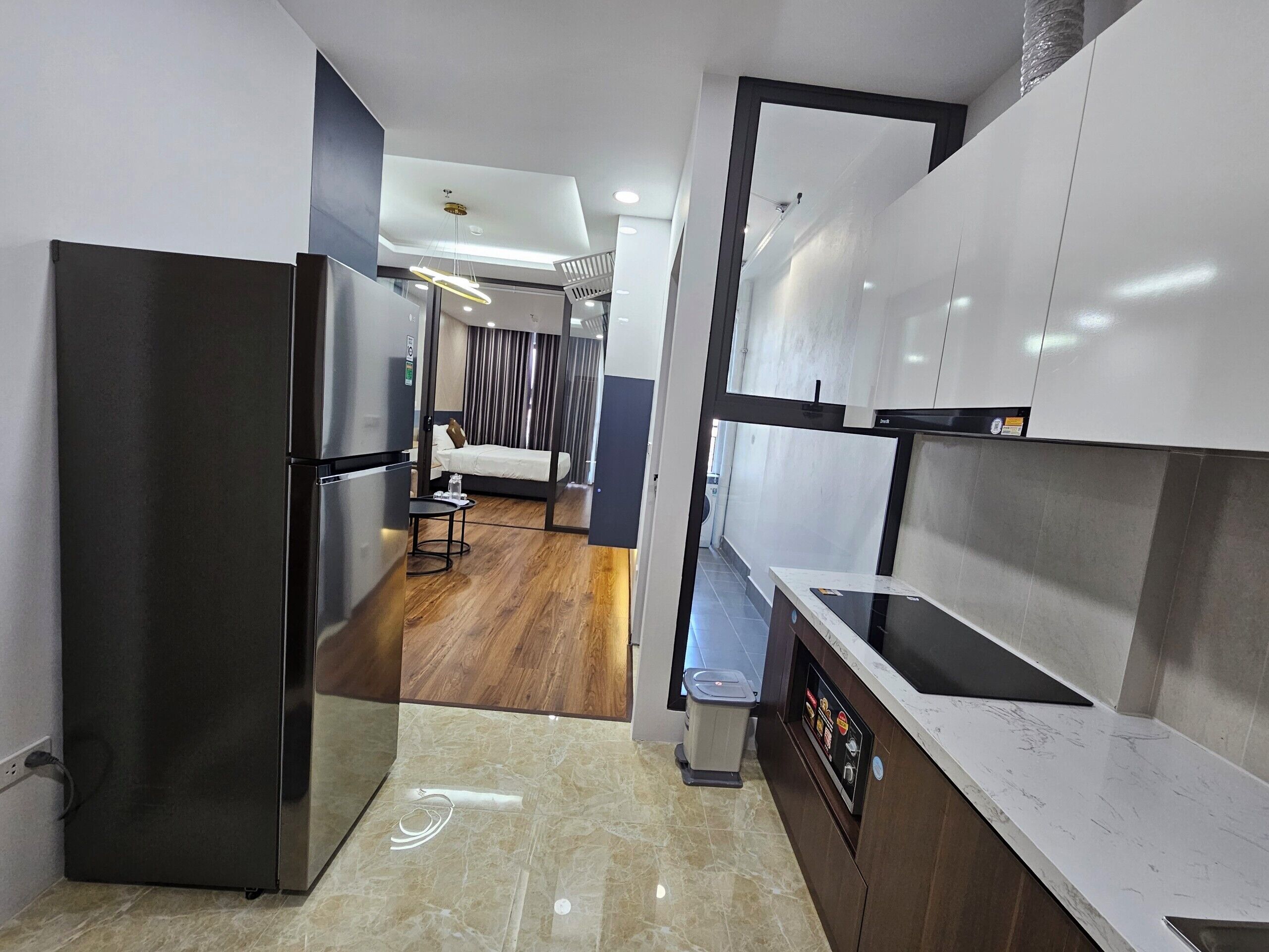 A modern kitchen with a built-in oven, a small dining area with a table and chairs, and a view into a bedroom with a bed and curtains.