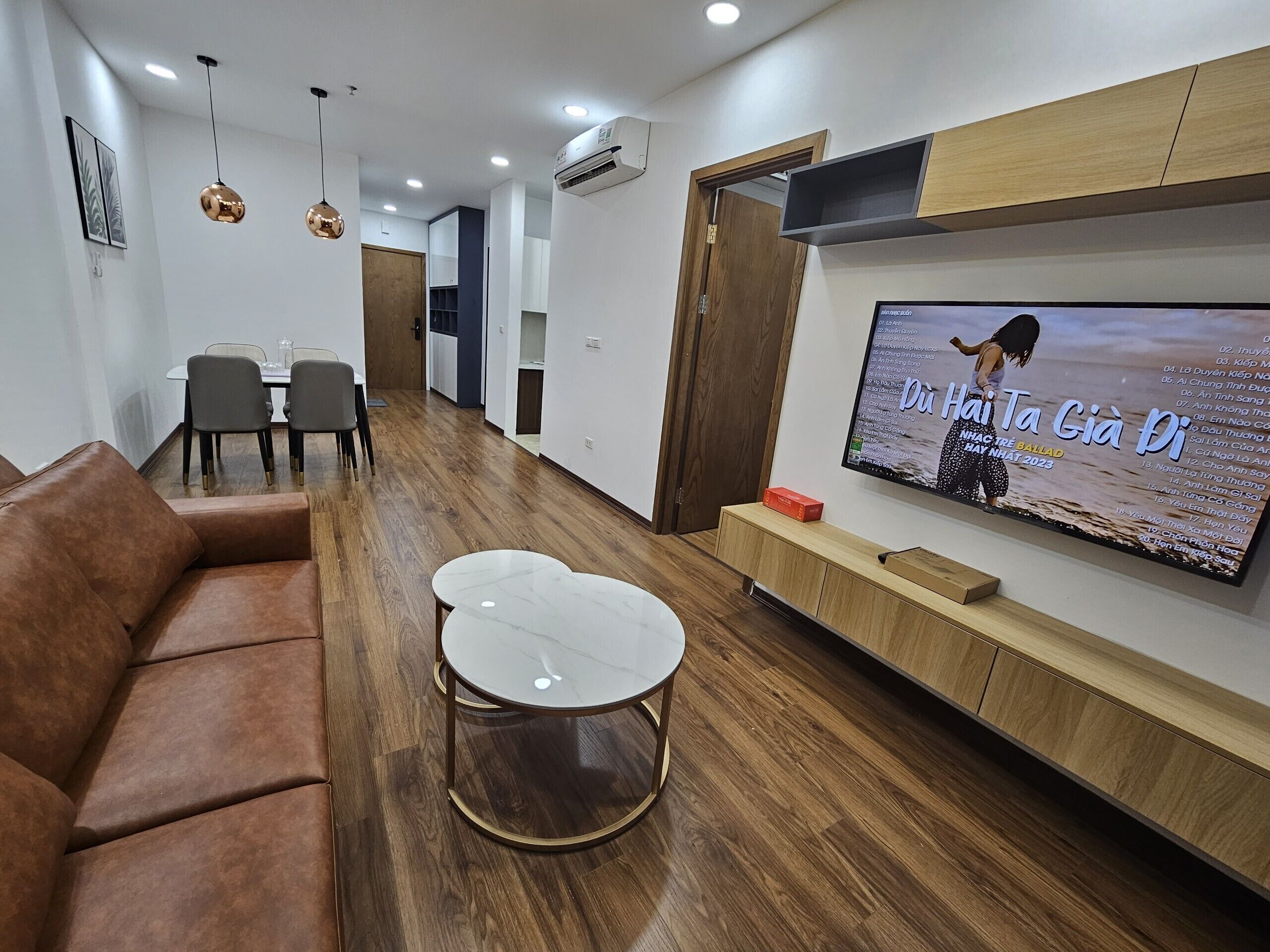 A modern living room with a brown leather sofa, a round coffee table, and a flat-screen TV mounted on the wall displaying a music video.