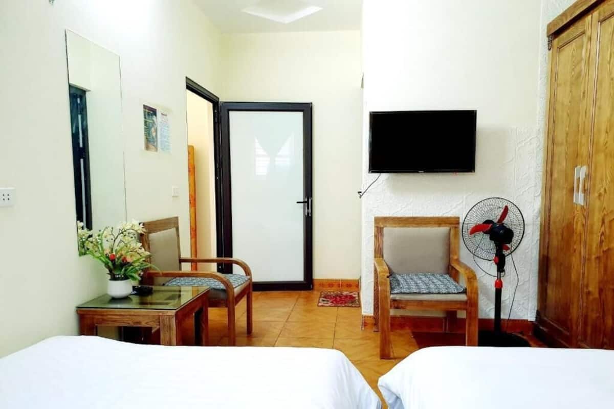 A hotel room with two beds, a desk with a plant, a chair, a wooden wardrobe, a TV, and a standing fan.