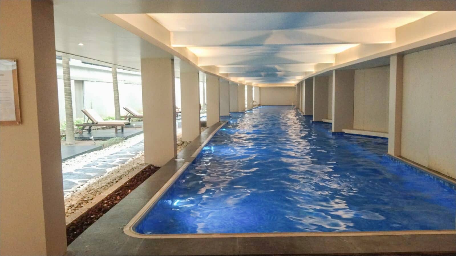A long indoor swimming pool with clear water, surrounded by walls and columns, and a bench area outside.