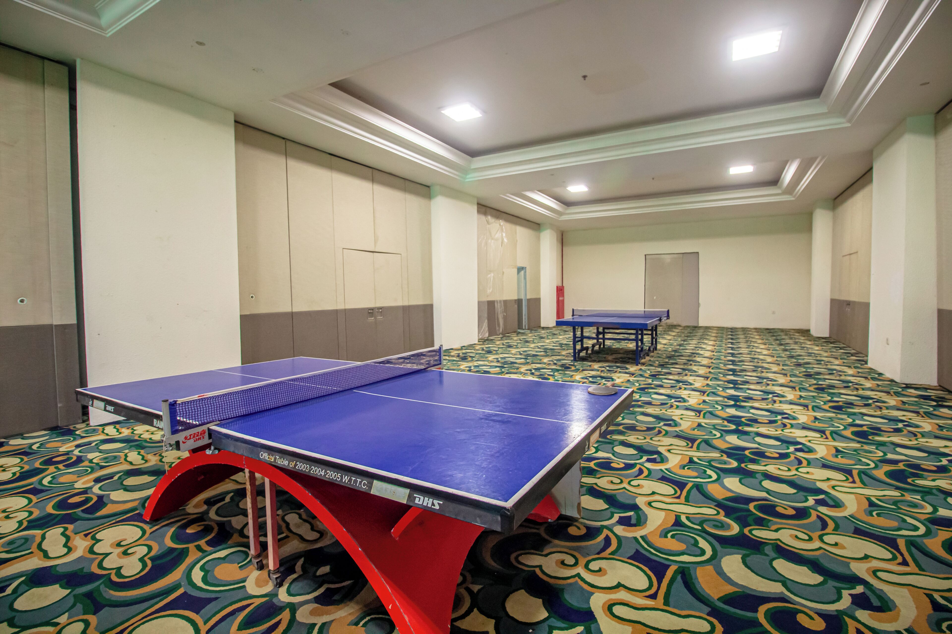 A room with a ping pong table and another table in the background, carpeted floor, and a door at the end of the room.