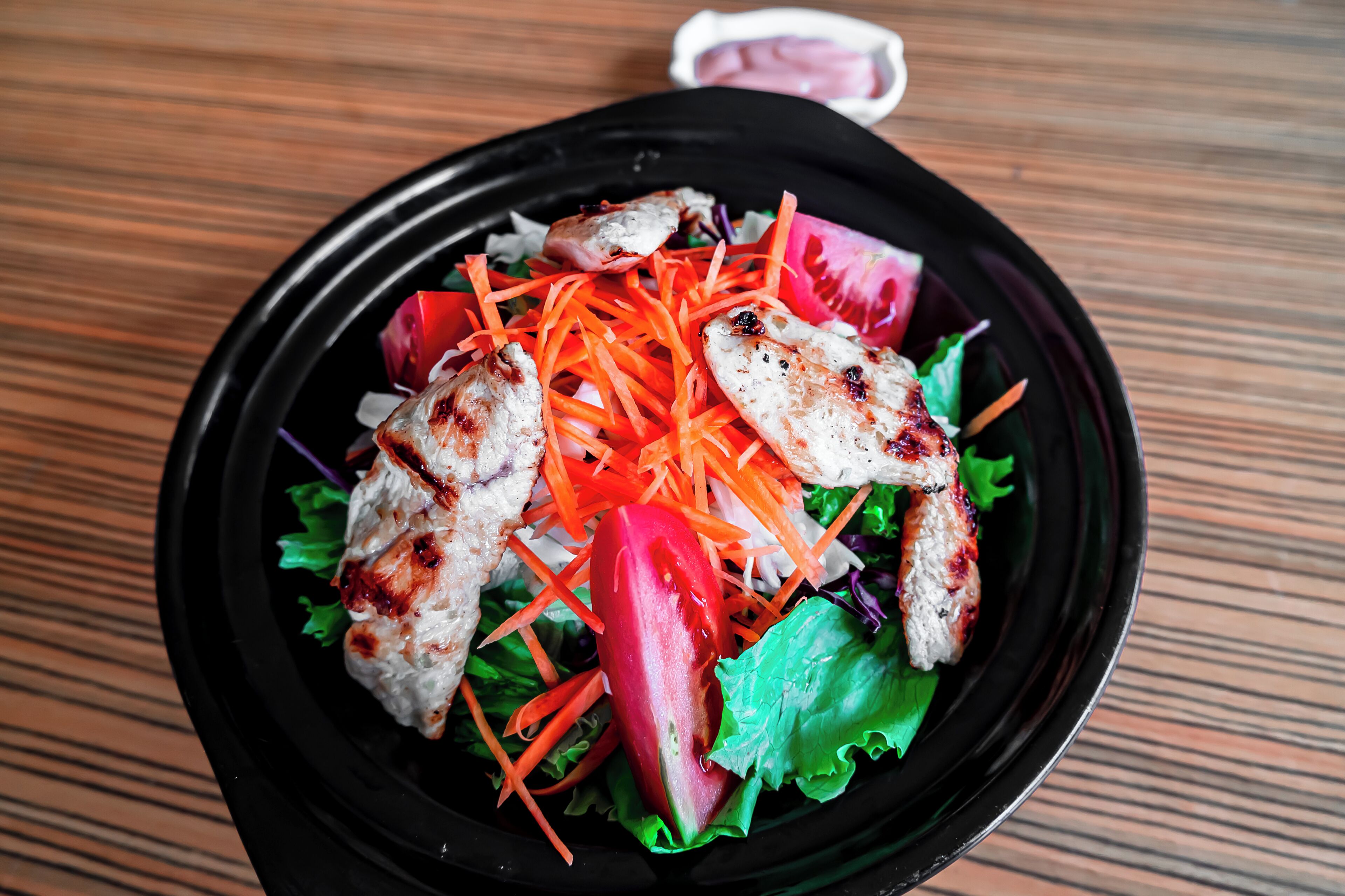 A salad with grilled chicken, shredded carrots, and lettuce in a black bowl on a wooden table.