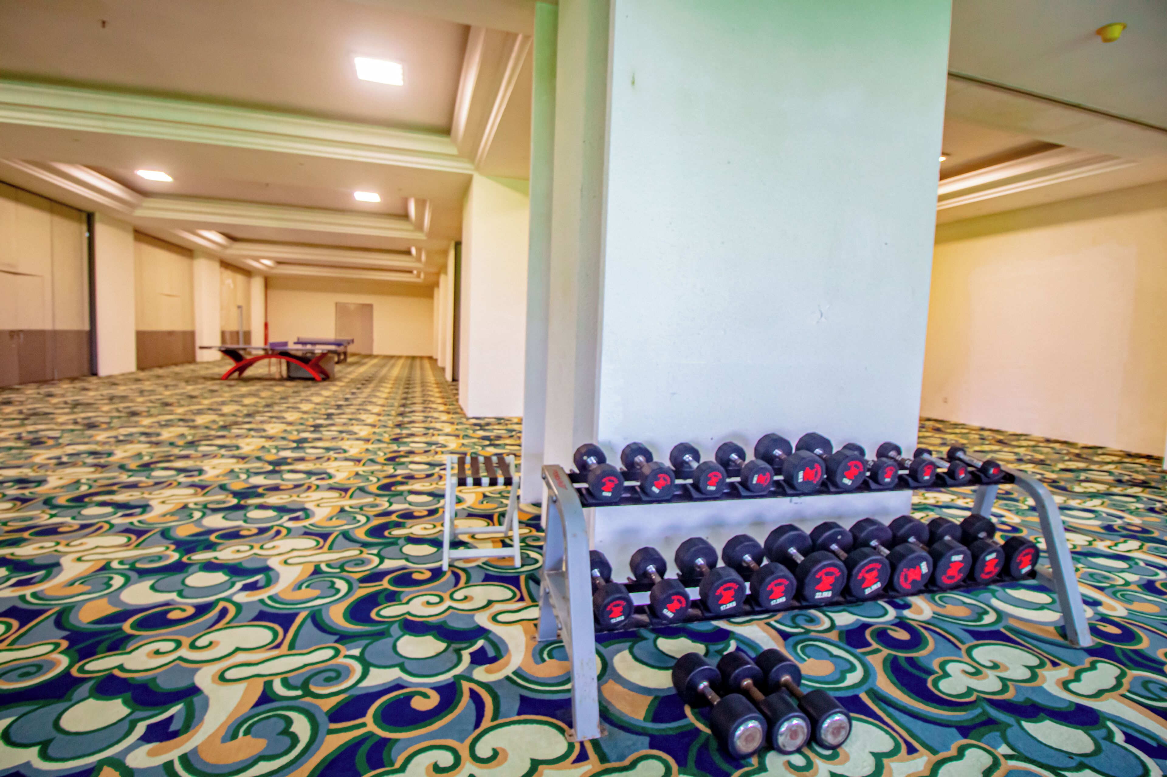 A hotel room with a patterned carpet, a table, and a rack of dumbbells.
