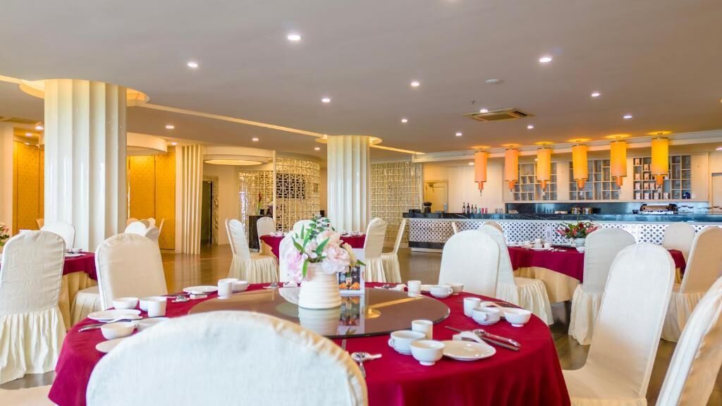 Een restaurantgedeelte in een hotel met ronde tafels gedekt voor een maaltijd, witte stoelen en een bar op de achtergrond.