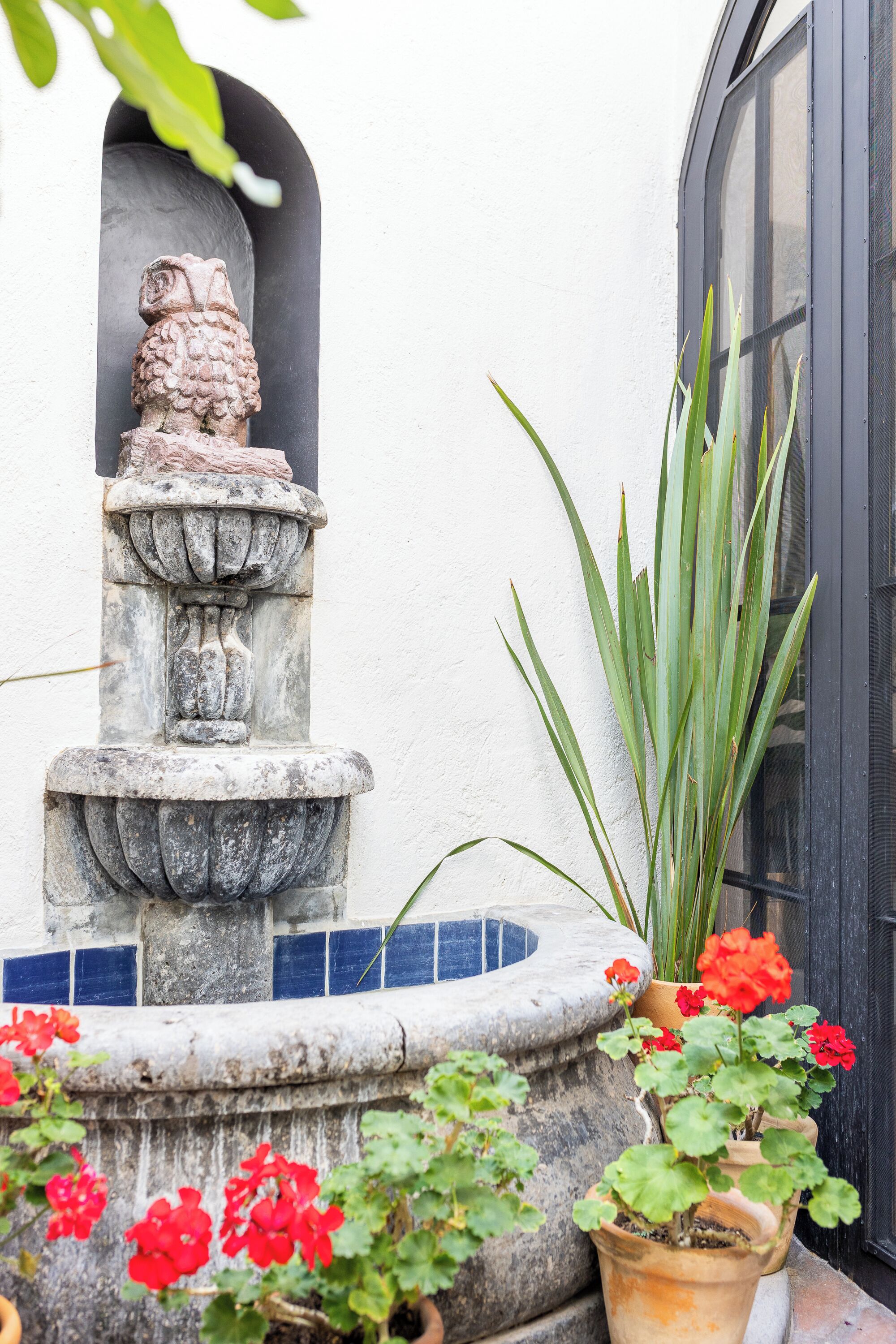 A stone fountain with a statue, surrounded by potted plants and red flowers.
