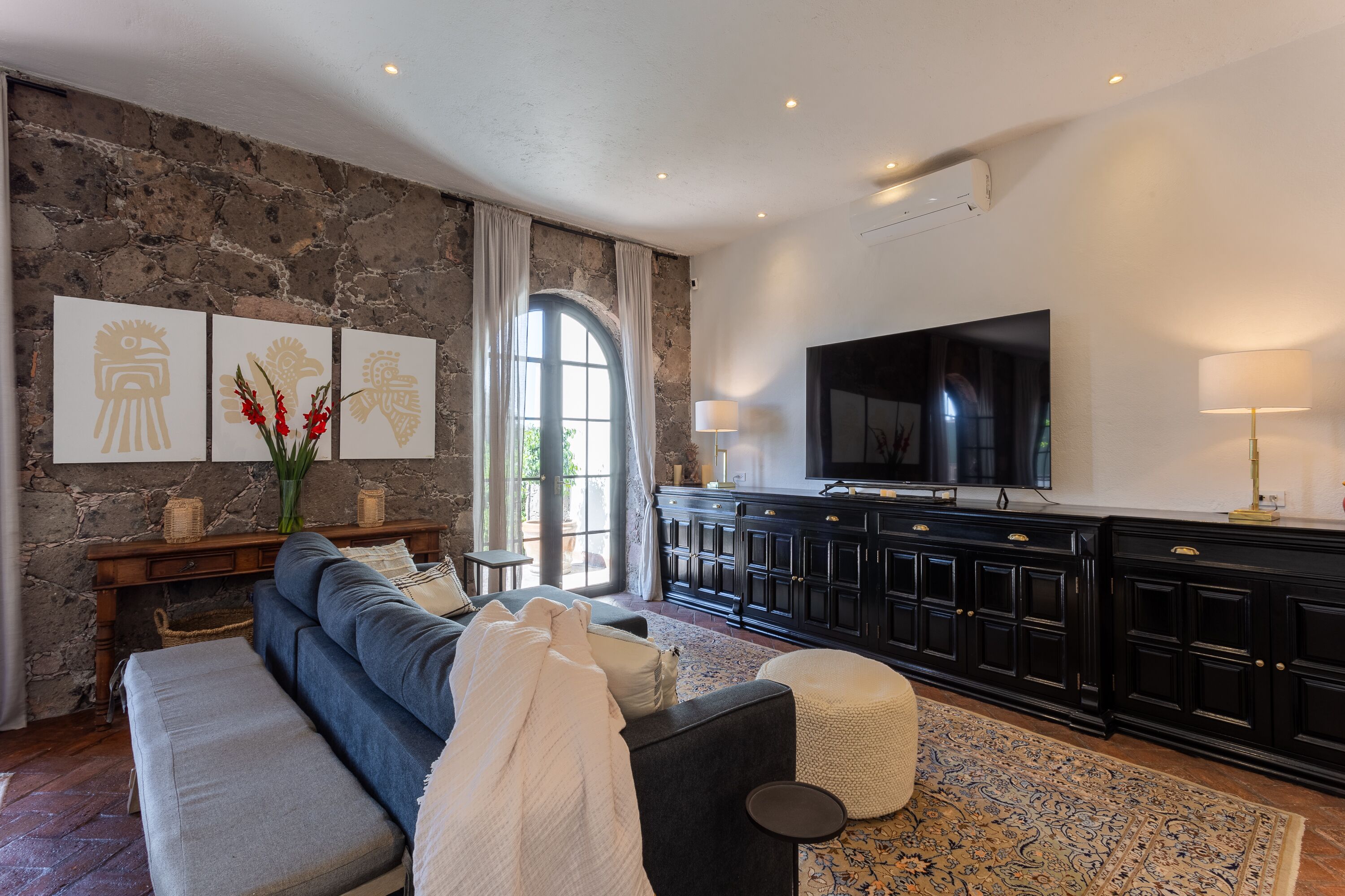 Una sala de estar con pared de piedra, un ventanal grande, un sofá, un televisor y un mueble.