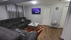 Una sala de estar con un sofá modular, una mesa de centro, un televisor de pantalla plana montado en la pared, una pequeña mesita auxiliar y una puerta con un reloj.