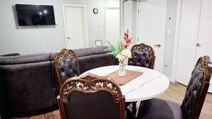 Un comedor con una mesa redonda, cuatro sillas tapizadas, un florero con flores y un televisor en la pared.