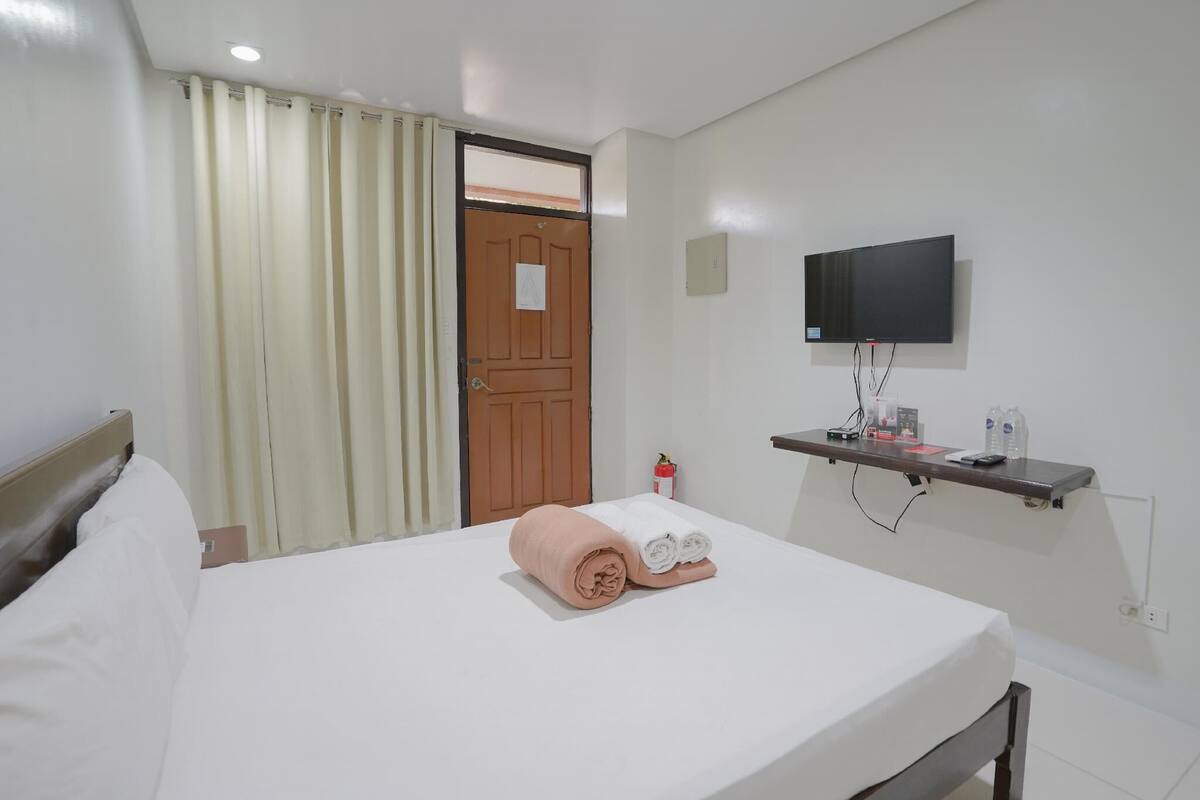 A hotel room with a bed, a television mounted on the wall, a small shelf with toiletries, and a door with a window.