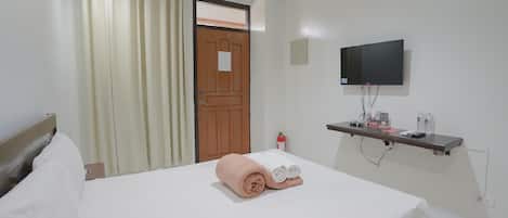 A hotel room with a bed, a television mounted on the wall, a small shelf with toiletries, and a door with a window.