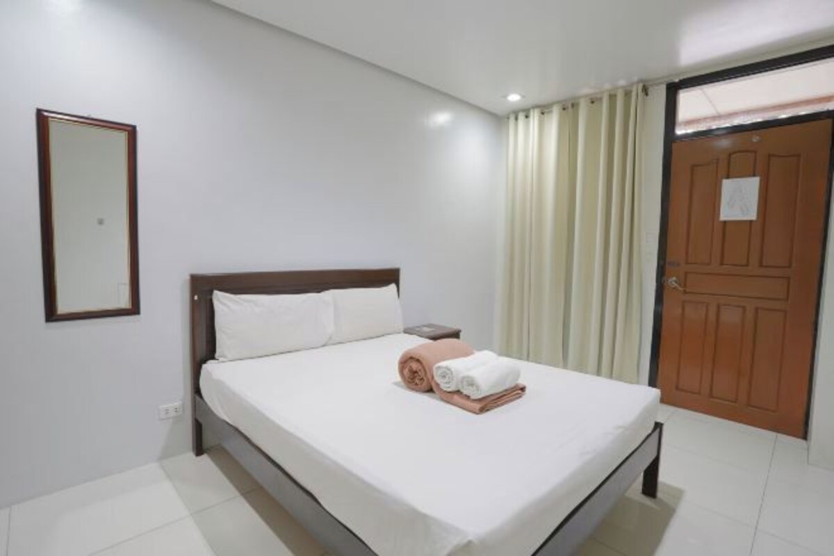 A neatly made bed with white linens, a wooden headboard, and a small bedside table. A mirror hangs on the wall to the left of the bed. A door with a wooden frame is visible to the right.