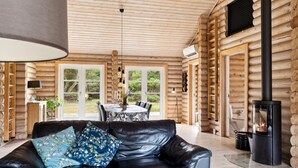 A log cabin interior with a leather sofa, dining table, and a wood-burning stove.