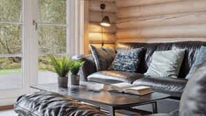 A cozy cabin interior with a leather sofa, a coffee table with books and a plant, and a large window offering a view of trees.