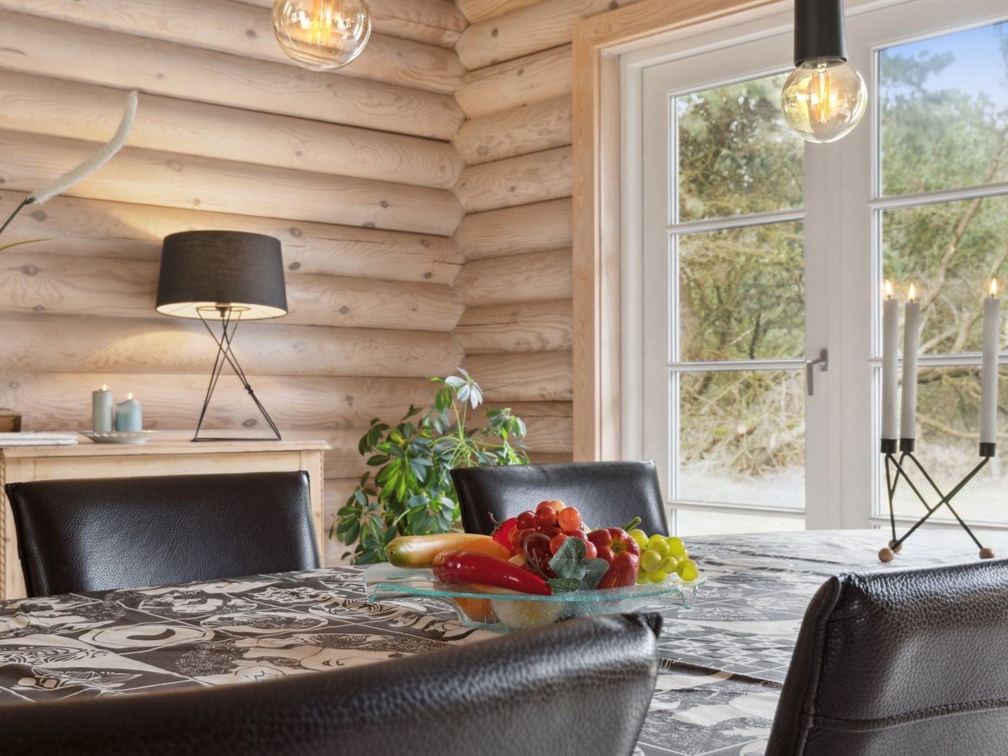 L’intérieur d’un chalet en bois, avec une table à manger, des chaises, une lampe et une vue sur l’extérieur.