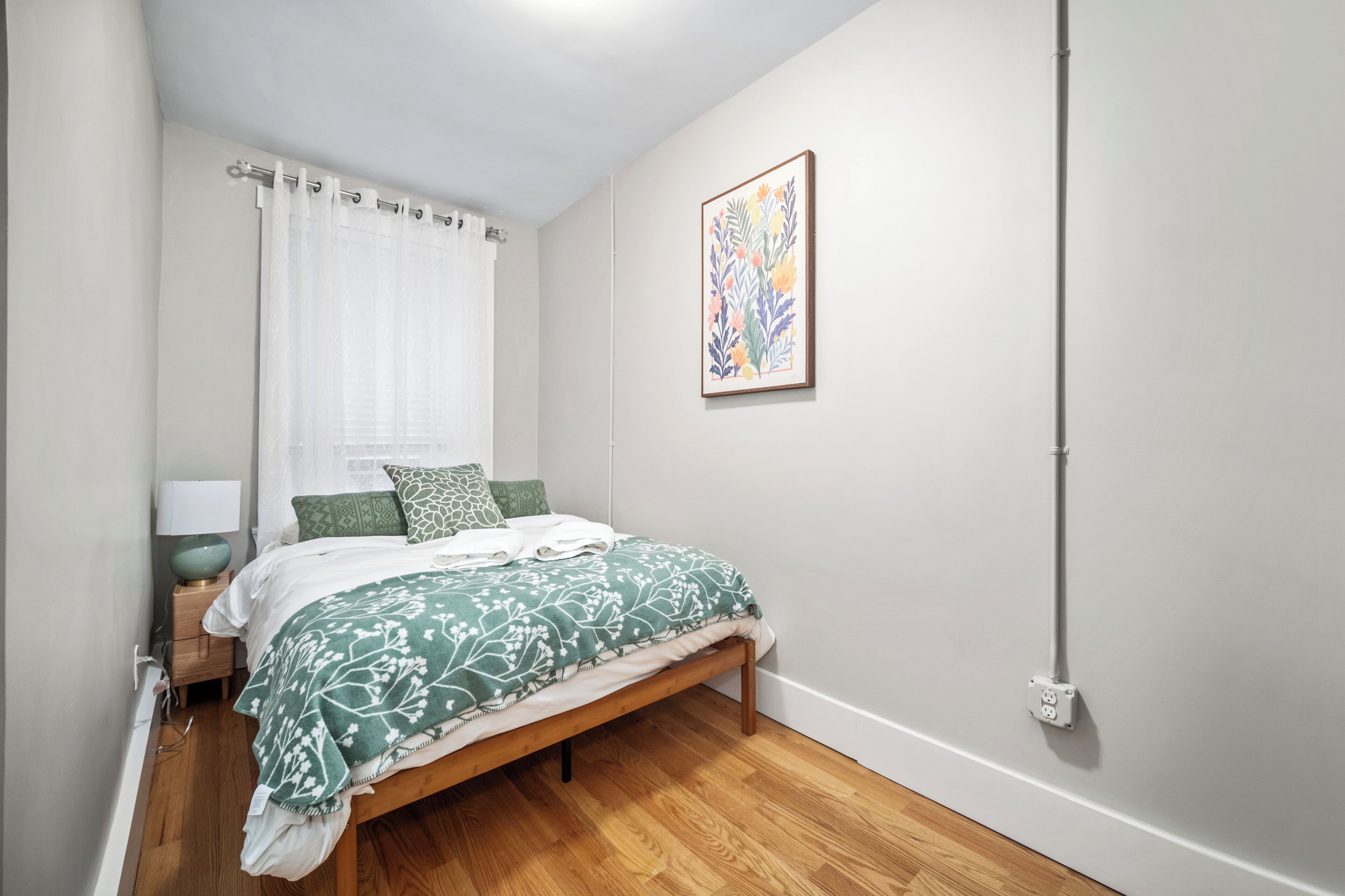 A bedroom with a wooden bed, white curtains, a green and white patterned bedspread, a green pillow, a bedside table with a lamp, and a framed artwork on the wall.
