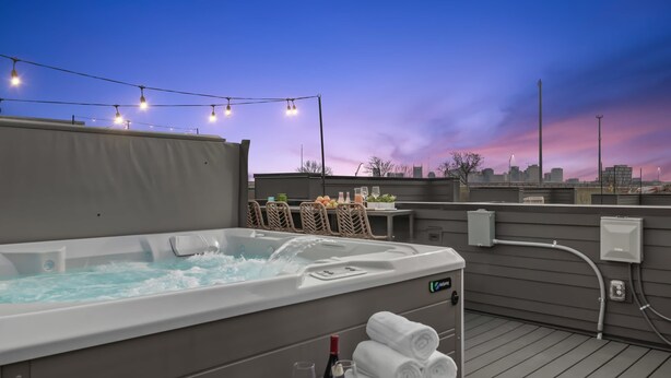 A hot tub with water running, surrounded by a deck with string lights, a table with chairs, and a city skyline in the background. - The Hubb House – Relaxing Rooftop + Hot Tub (Nashville)
