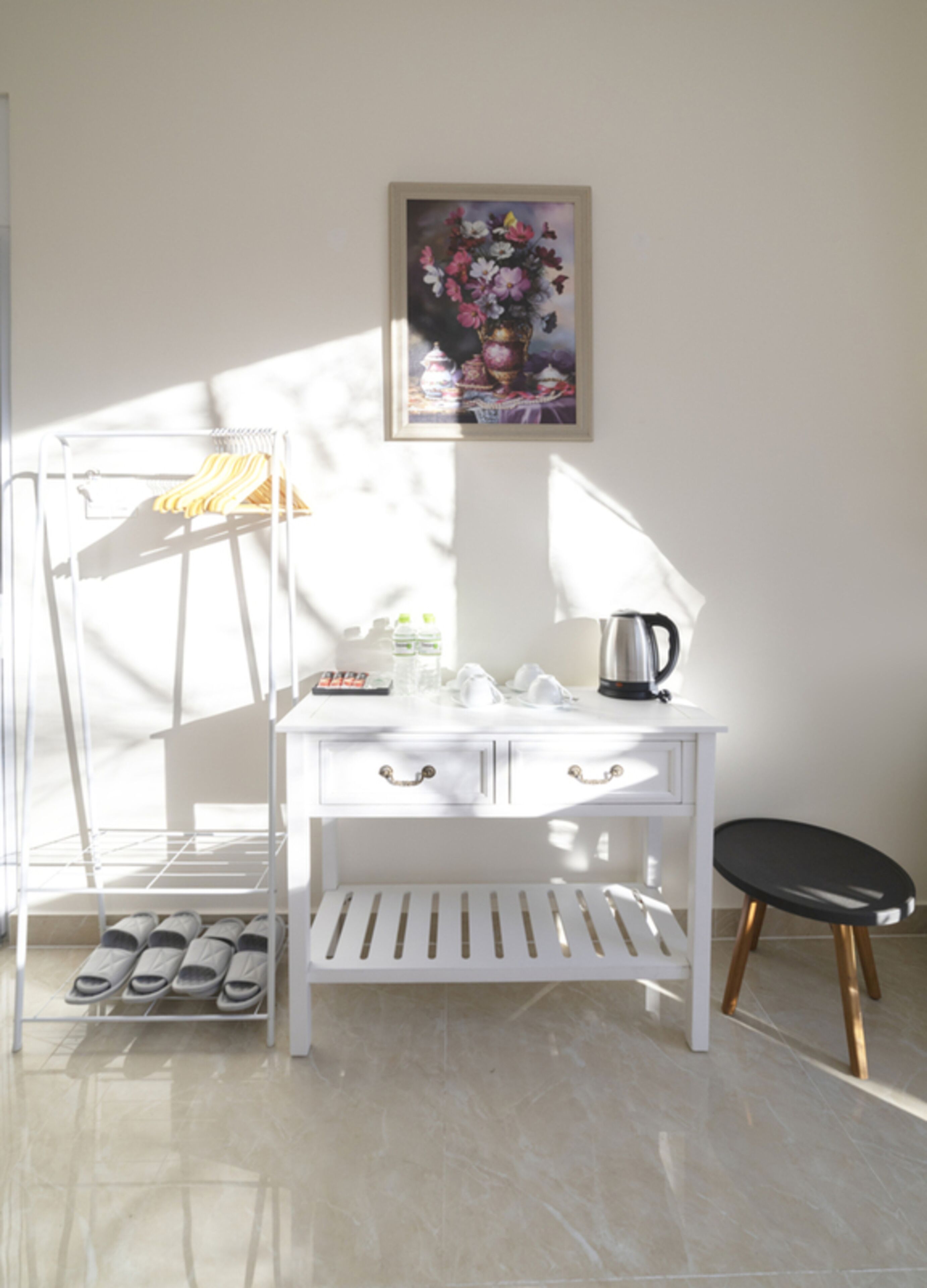 A white desk with drawers, a black stool, a shoe rack, and a framed floral painting on the wall.