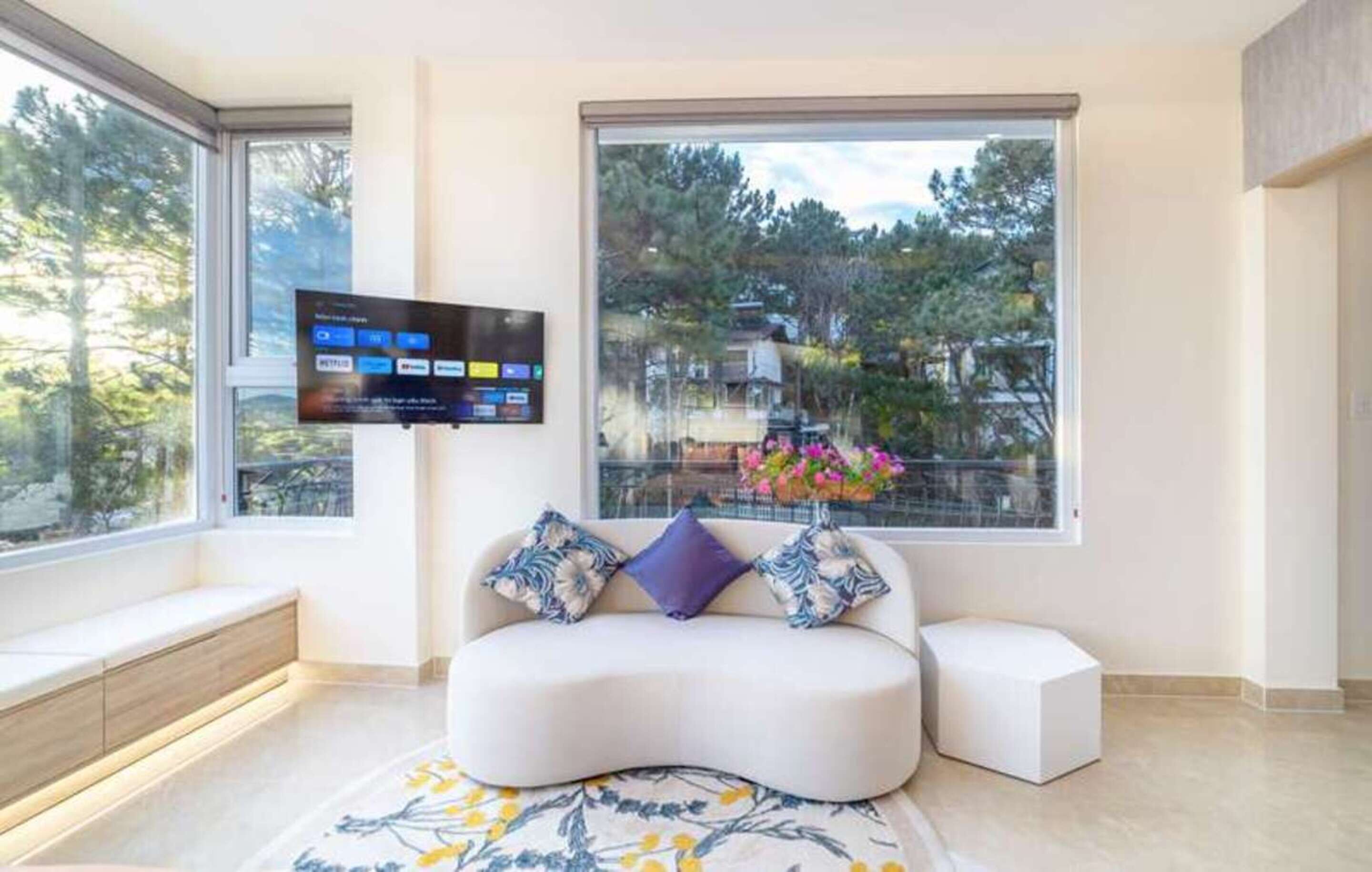 A modern living room with a sofa, a flat-screen TV mounted on the wall, a window with a view of trees, and a small white ottoman.