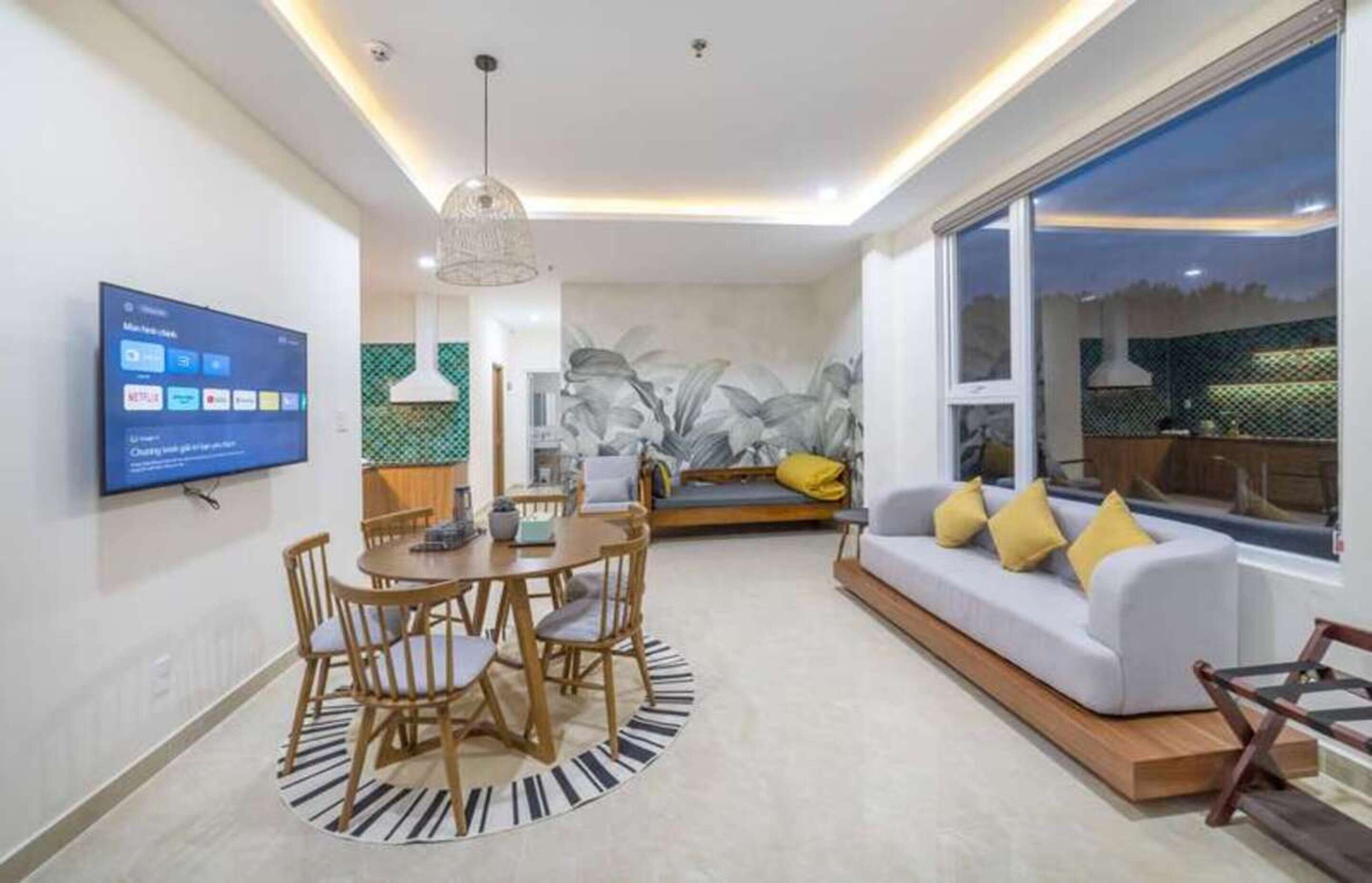 A modern living room with a flat-screen TV, a dining area with a round table and chairs, a sofa with yellow cushions, and a large window offering a view of the outdoors.